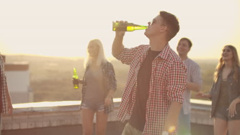 A-young-man-in-trendy-glasses-drinks-beer-and-moves-in-a-dance-at-a-party-with-his-friends-on-the-roof.